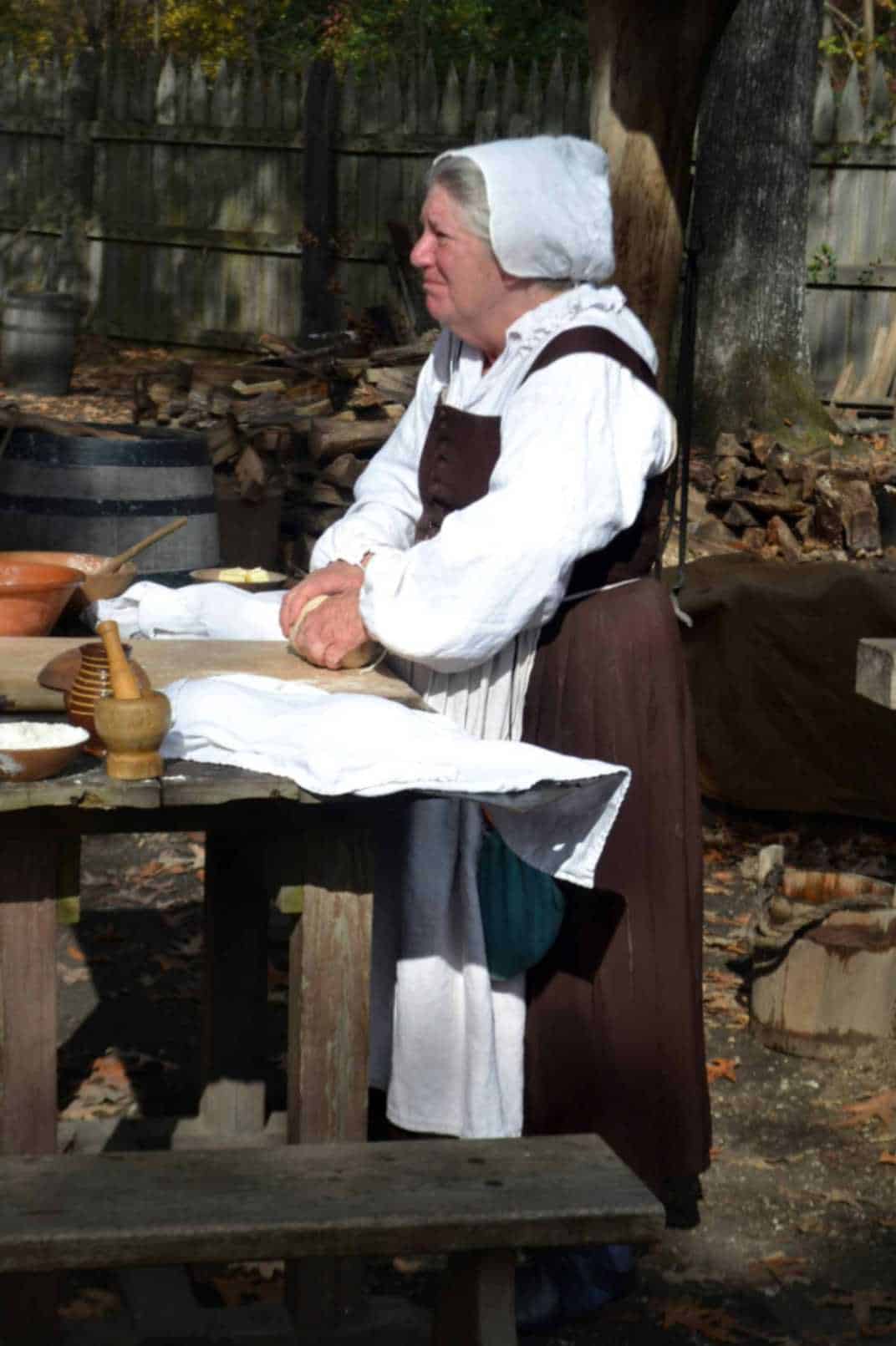 An old woman dressed in 18th-century maid clothes making food outdoors.