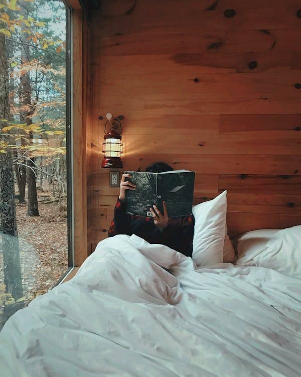 A person inside a cabin, lying down on a white bed while holding a book.