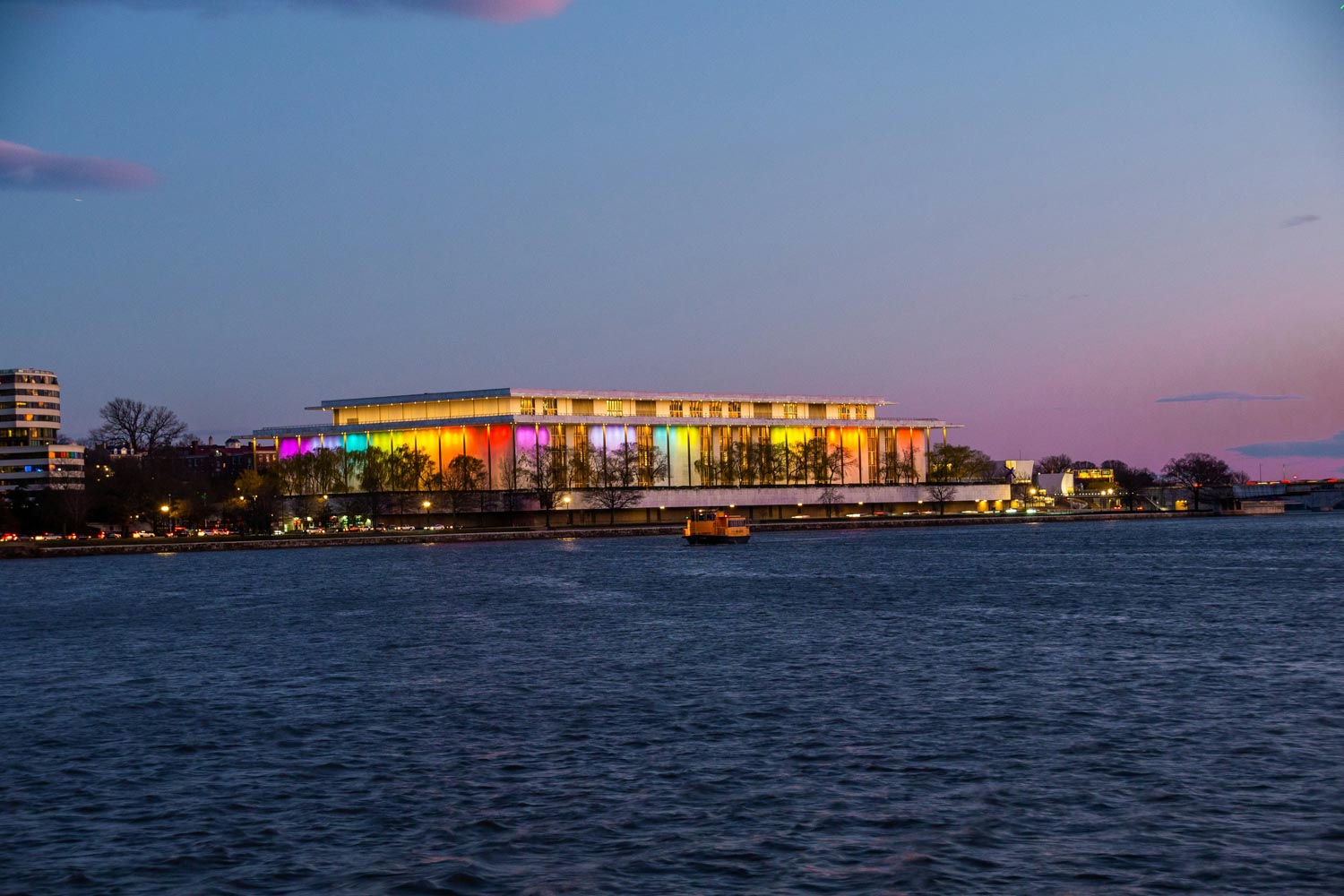 A view of a building in Washington DC with colorful lighting seen from across a body of water at night.