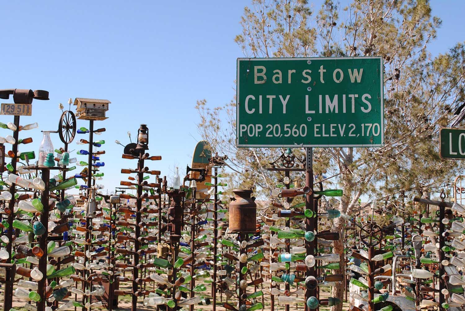 Art installations of old tools, mostly bottles, in a desert, with a sign "Barstow City Limits".