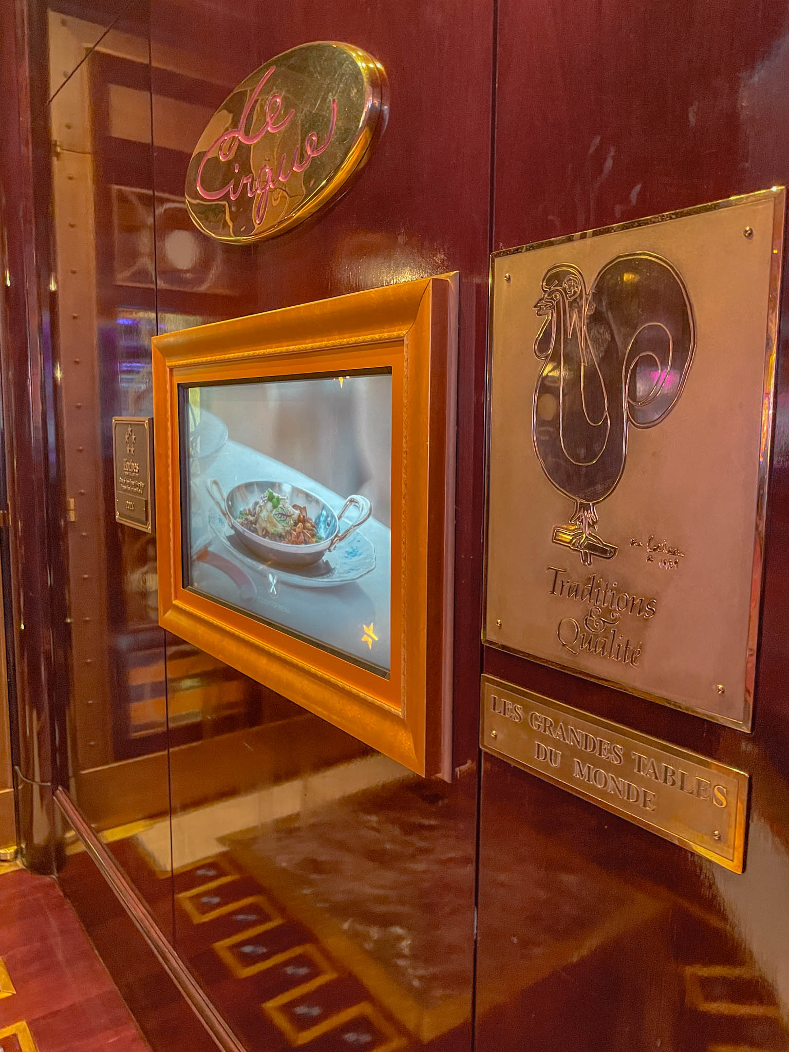 The wall of a restaurant with golden carving of name and a figure of a chicken, and a screen showing food.