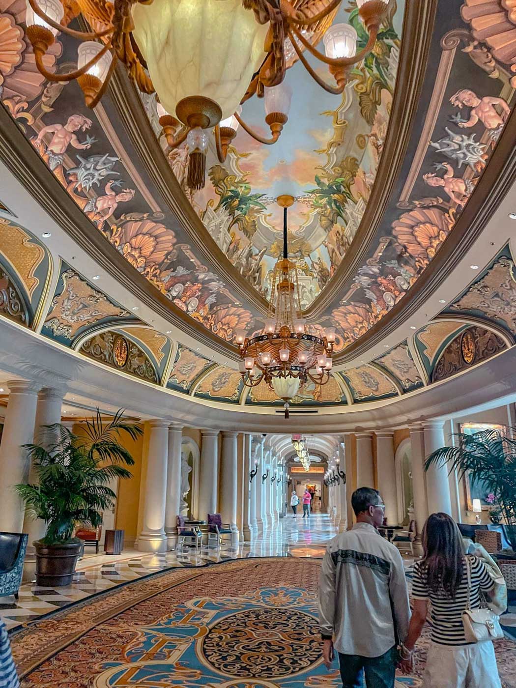 People walking on the hallways of a hotel with colorful flooring and carpets and painted ceilings.