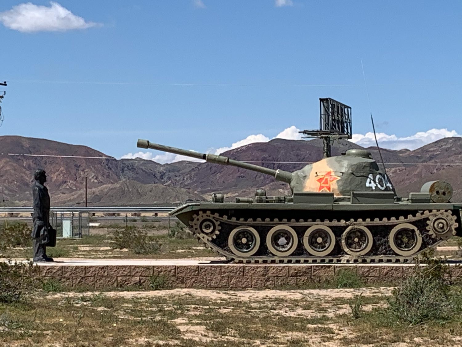 A sculpture park with figures of a human and tank in a desert with rocky mountain views in the background.