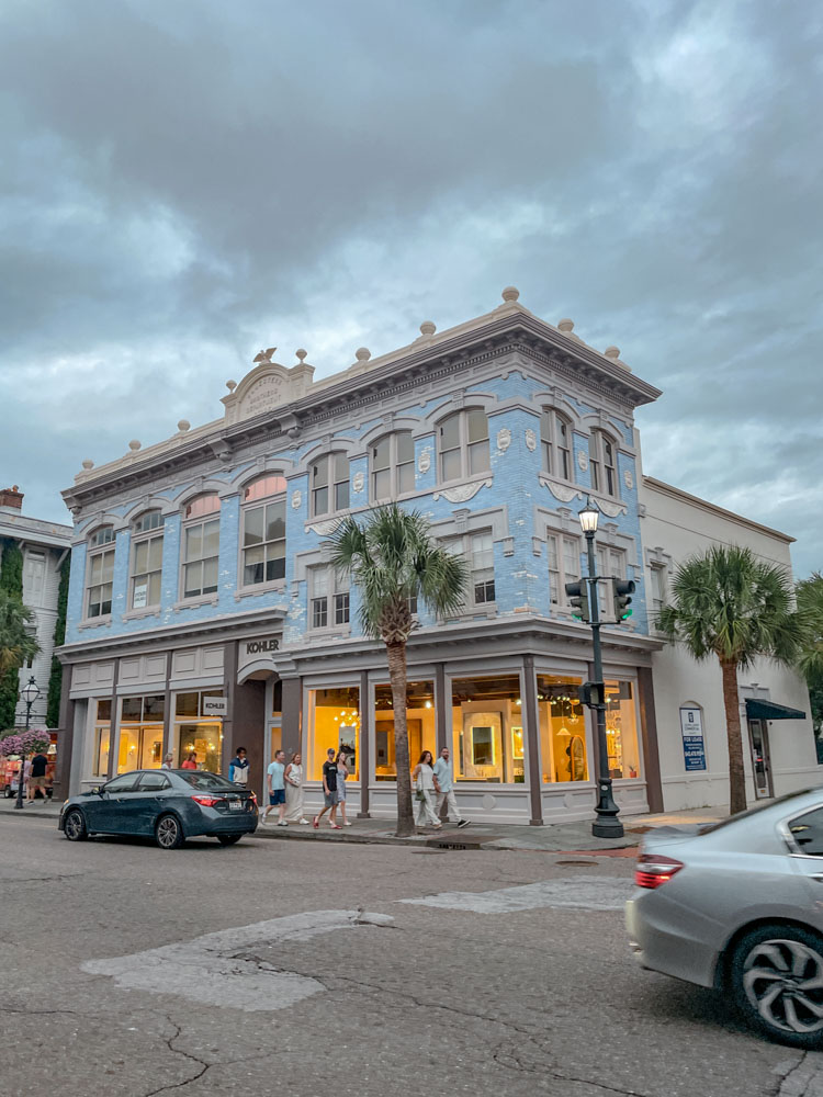 Cars passing by, people walking on a side walk in front of a cute building in Charleston, South Carolina.