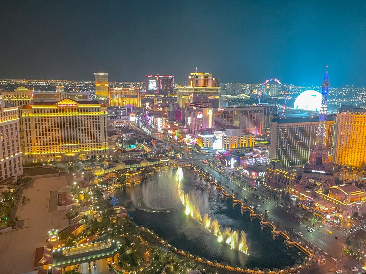 An aerial view of Las Vegas at night with hotels, roller coaster, fountains, and mini Eiffel Tower.