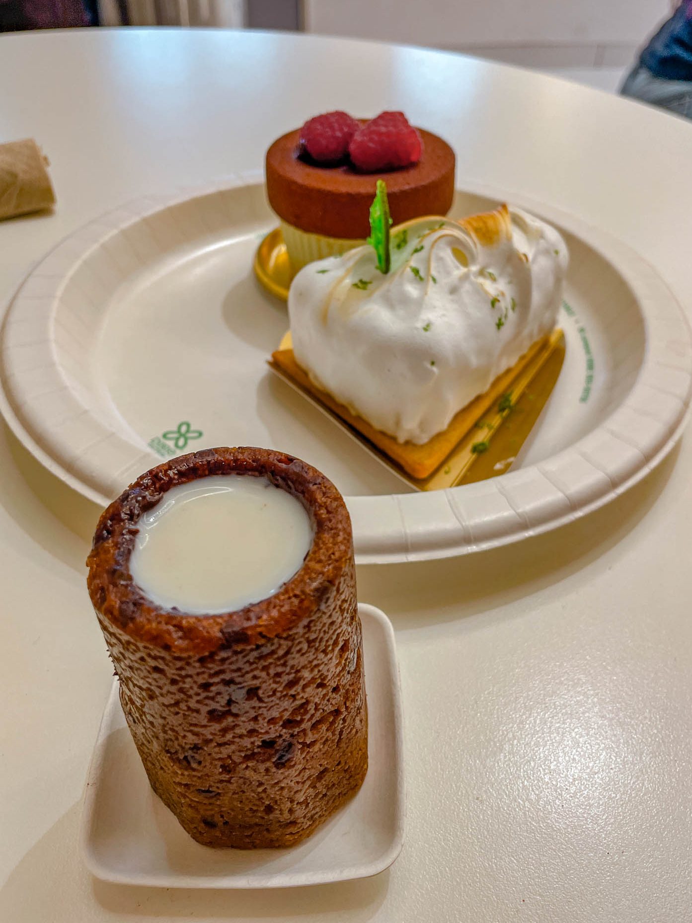 Cakes and tart on a paper plate and a cookie shaped like a cup with milk.