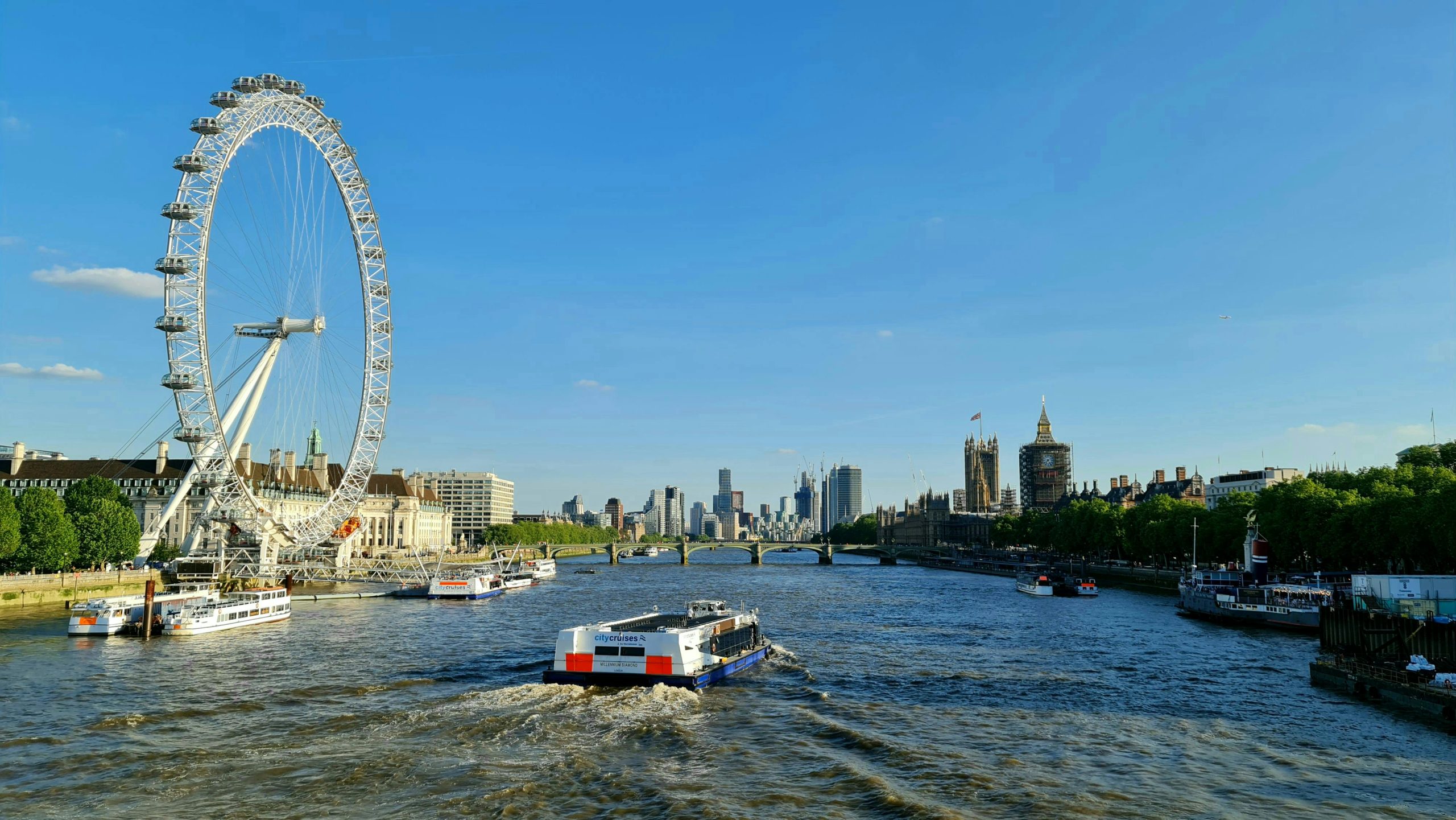 A cruise boat on the River Thames in London, England, with the view of the city buildings and the London Eye ferris wheel.