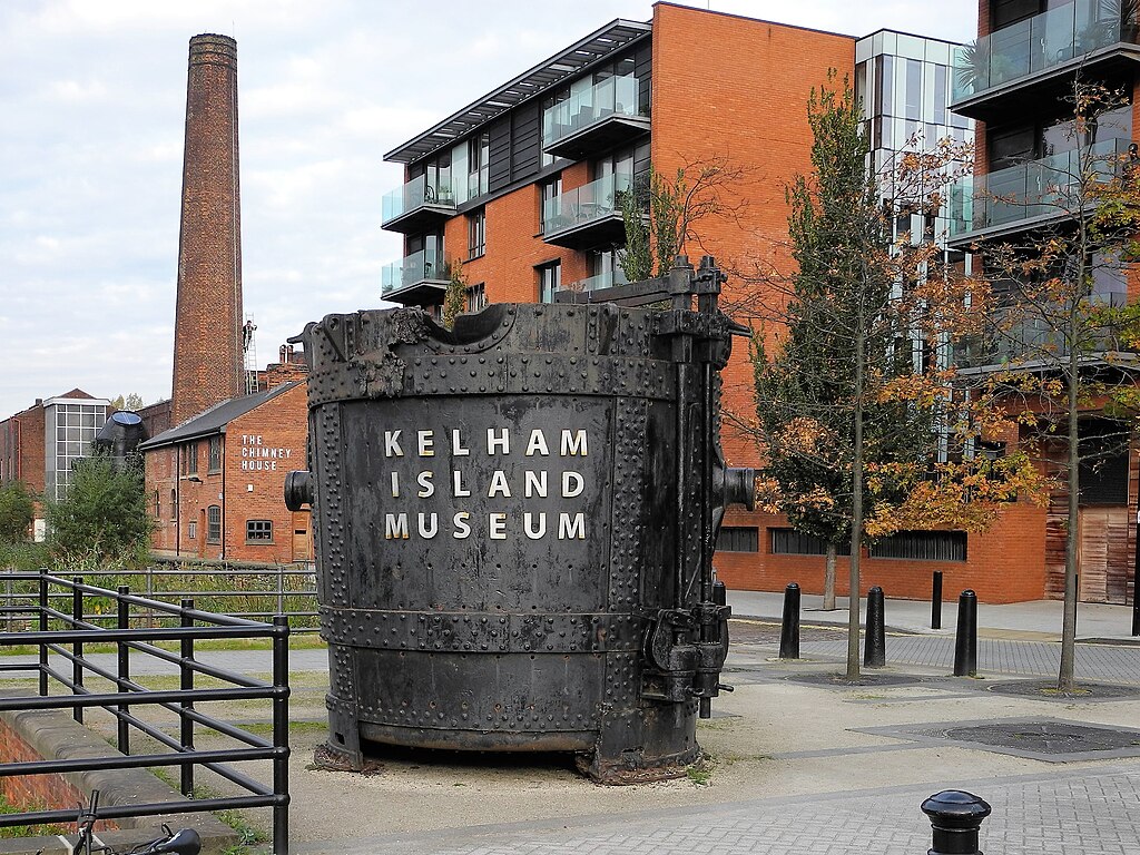 Giant steel ladle with text "Kelham Island Museum" displayed outdoors surrounded by orange buildings and trees in Sheffield, UK.