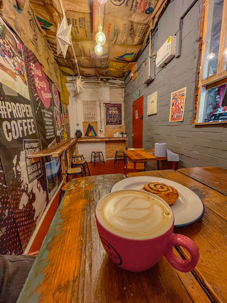 Latte on a red mug and a plate of pastry resting on a wooden table inside a small coffee shop in Sheffield, UK.