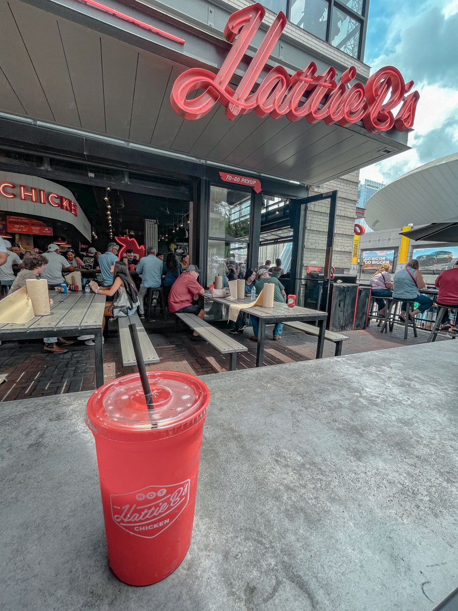 A cup of drink sitting on a table with the storefront and al fresco dining area of Hattie B's in Nashville Tennessee in the background.