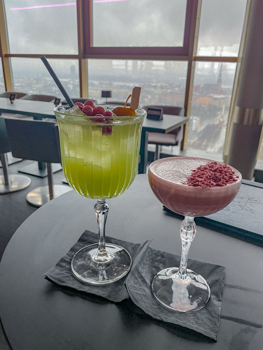Fancy drinks on a black round table at a hotel bar on its top floor, with a view of buildings outside from the window in the background.