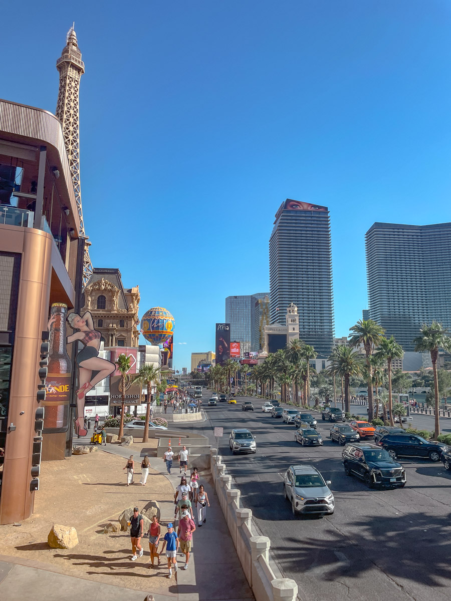 People walking on the side of the road and cars on the road with buildings and palm trees in the background in Las Vegas.
