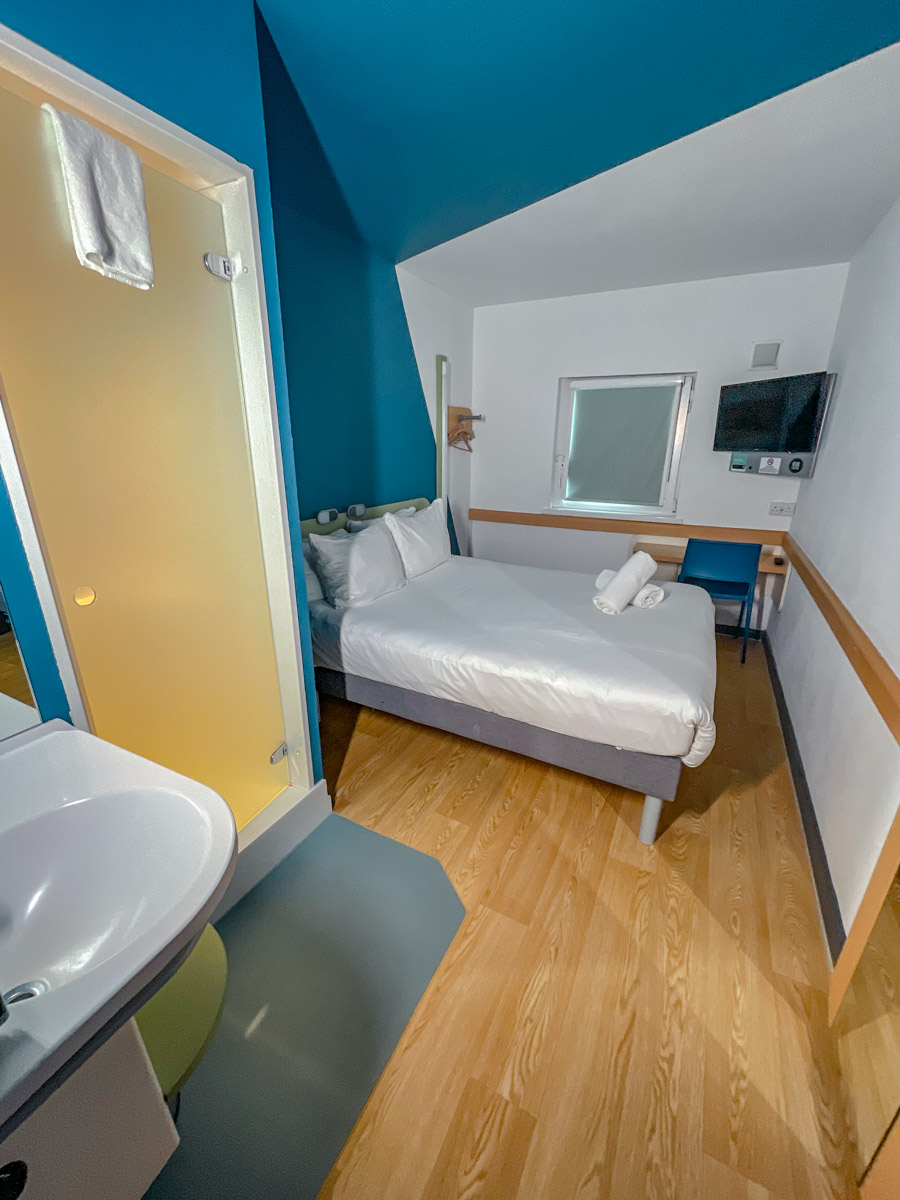 A hotel room with bed, table, chair, television, and sink.