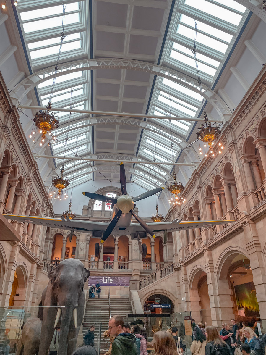 People inside Kelvingrove Art Gallery with replicas of old airplanes and of an elephant.