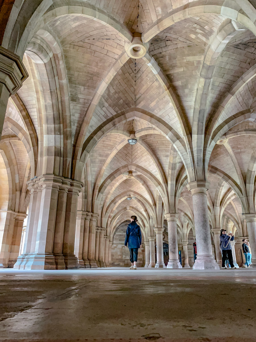 A woman standing under Gothic Revival vaulted stone ceilings and fluted columns at the University of Glasgow.