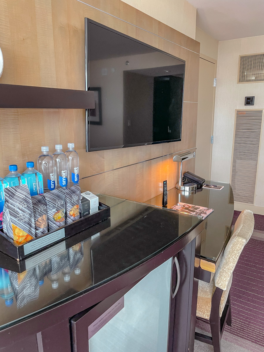 Hotel counter with supplies of food and bottles of water and a television mounted on a wall.
