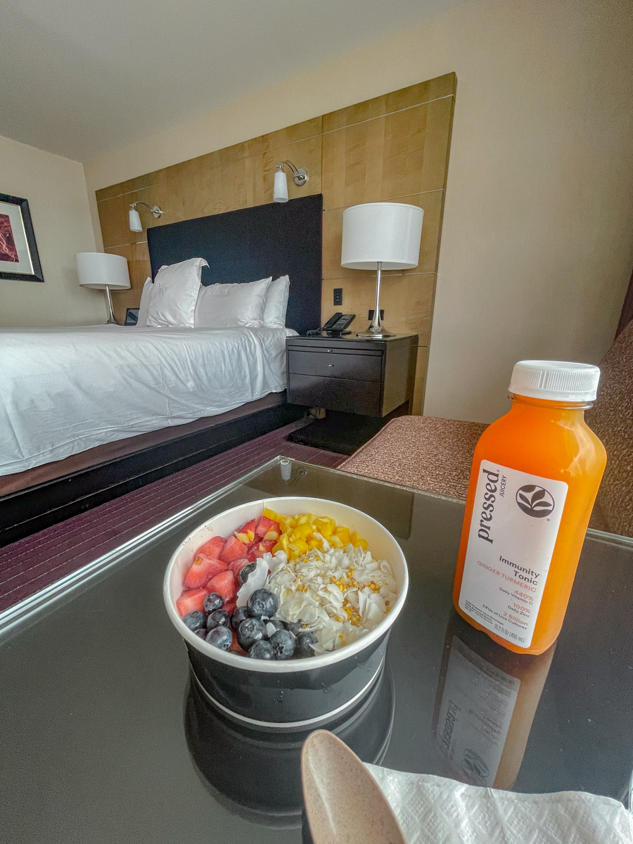 A juice and a bowl of fruits sitting on a hotel room table with a bed in the background.